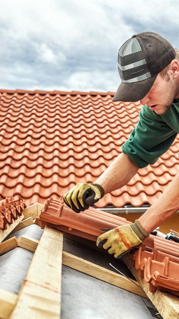 Un couvreur, portant des gants et une casquette, pose des tuiles en terre cuite.