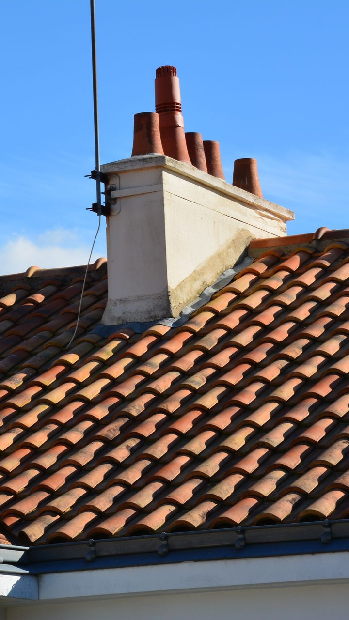 Toit de tuiles rouges avec cheminée et antenne sur fond de ciel bleu.