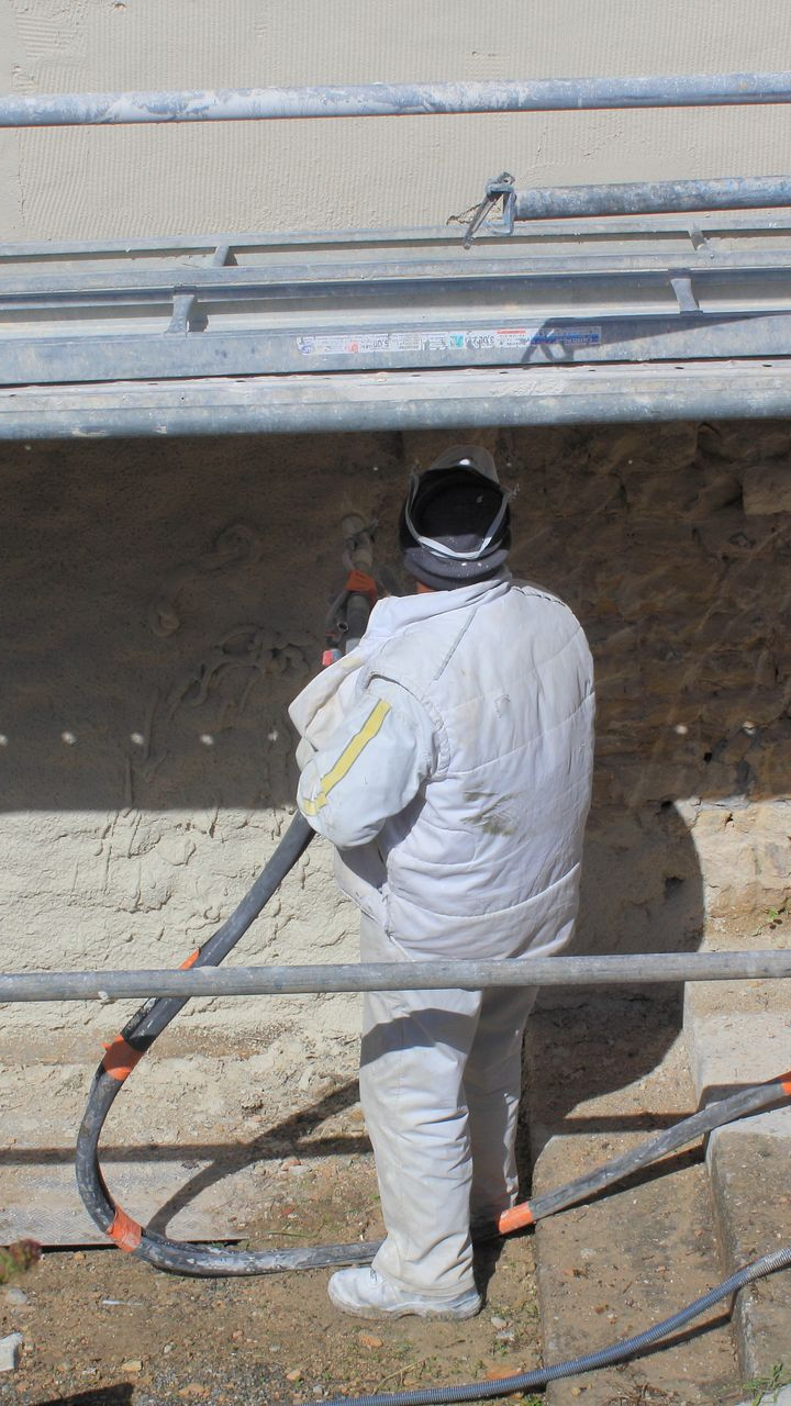Ouvrier en combinaison de protection, utilisant une buse pour traiter un mur, près d'un échafaudage.