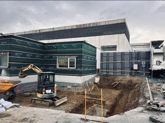 Chantier de construction avec une excavatrice à côté d'un bâtiment moderne partiellement construit et d'une excavation de terre.