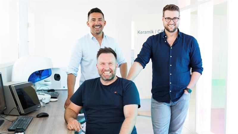 Drei Männer stehen nebeneinander vor einem Schreibtisch.