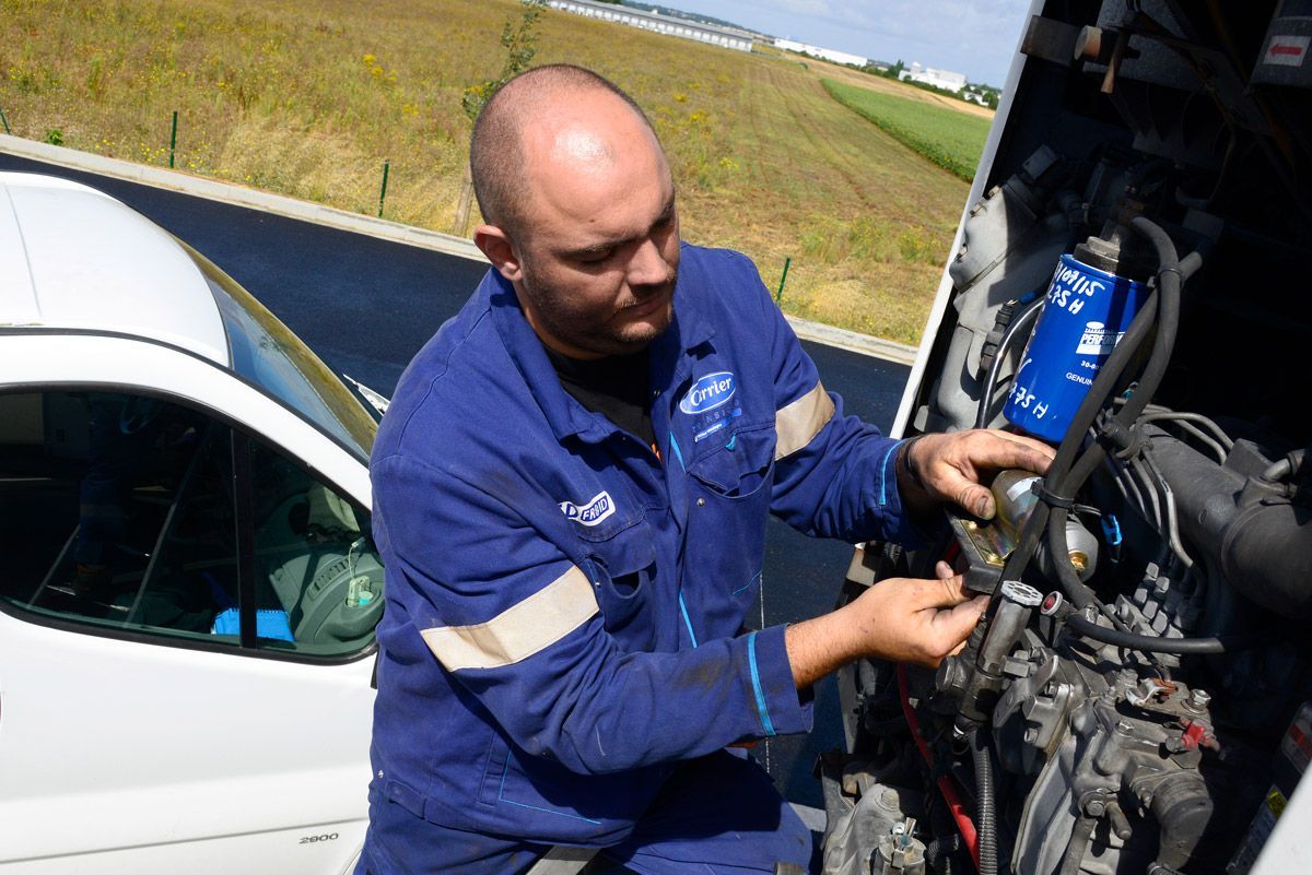 Un mécanicien en combinaison bleue travaille sur le moteur d'une voiture à découvert, à l'extérieur, à côté d'un véhicule blanc