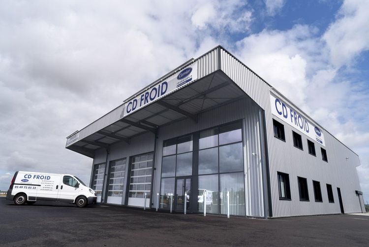 Bâtiment commercial moderne blanc avec une grande entrée vitrée et une camionnette blanche garée à l'extérieur sous un ciel nuageux