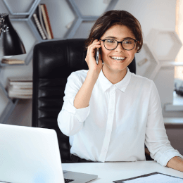 Eine Frau mit Brille sitzt an einem Schreibtisch und telefoniert mit dem Handy