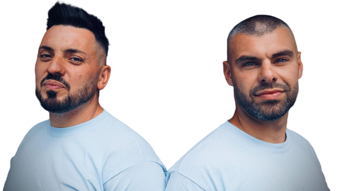 Zwei Männer posieren Rücken an Rücken und blicken in die Kamera. Einer hat eine Tolle, der andere einen rasierten Kopf.
