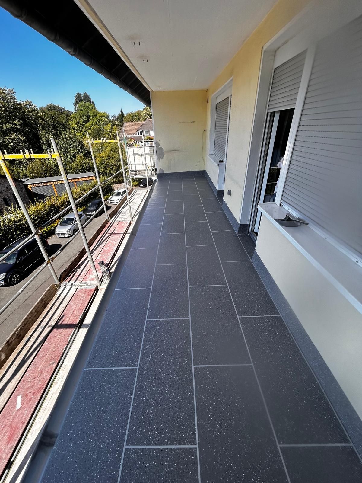 Schmaler Balkon mit dunkelgrauen Fliesen, außen ein Gerüst, gelbe Wände und geschlossene graue Fensterläden.