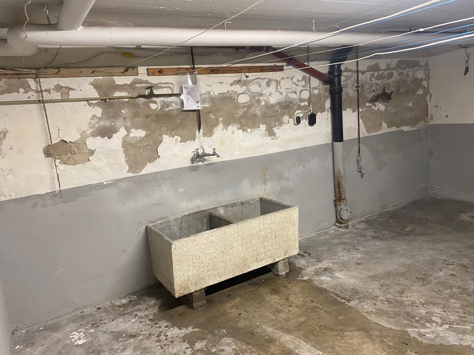 Betonwaschbecken in einem feuchten Keller mit abblätternden Wänden und freiliegenden Rohren.