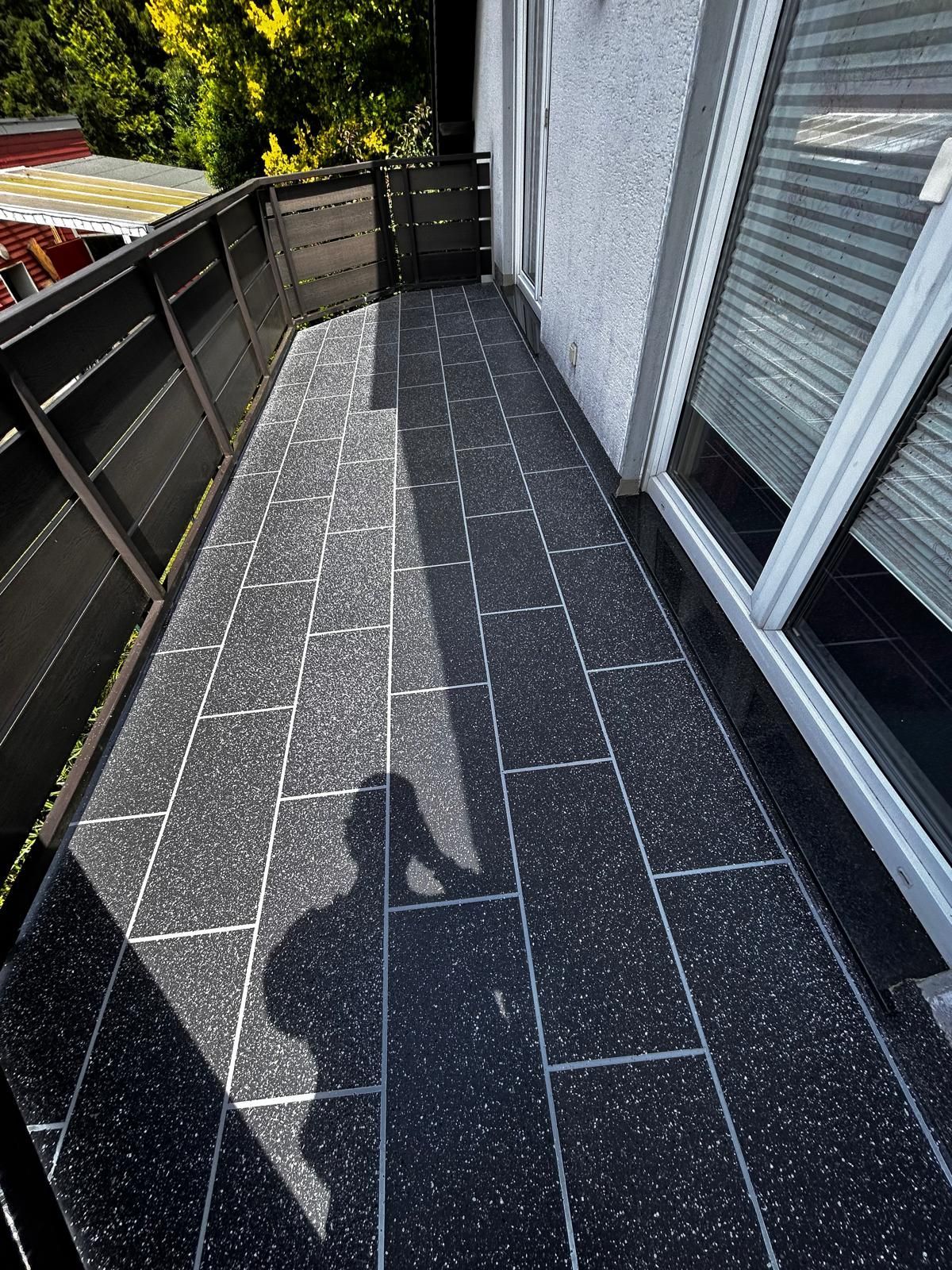 Balkon mit dunkel gesprenkelten Fliesen, schwarzem Geländer und Schatten einer Person.