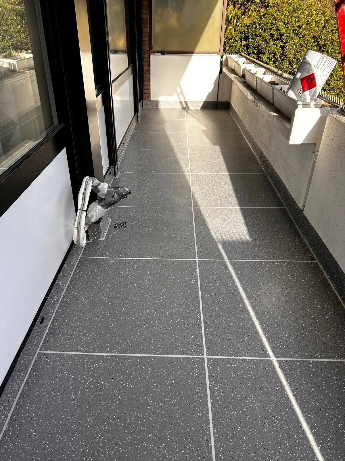 Schmaler Balkon mit grau gesprenkeltem Fliesenboden und weißen Wänden. Sonnenlicht wirft lange Schatten.