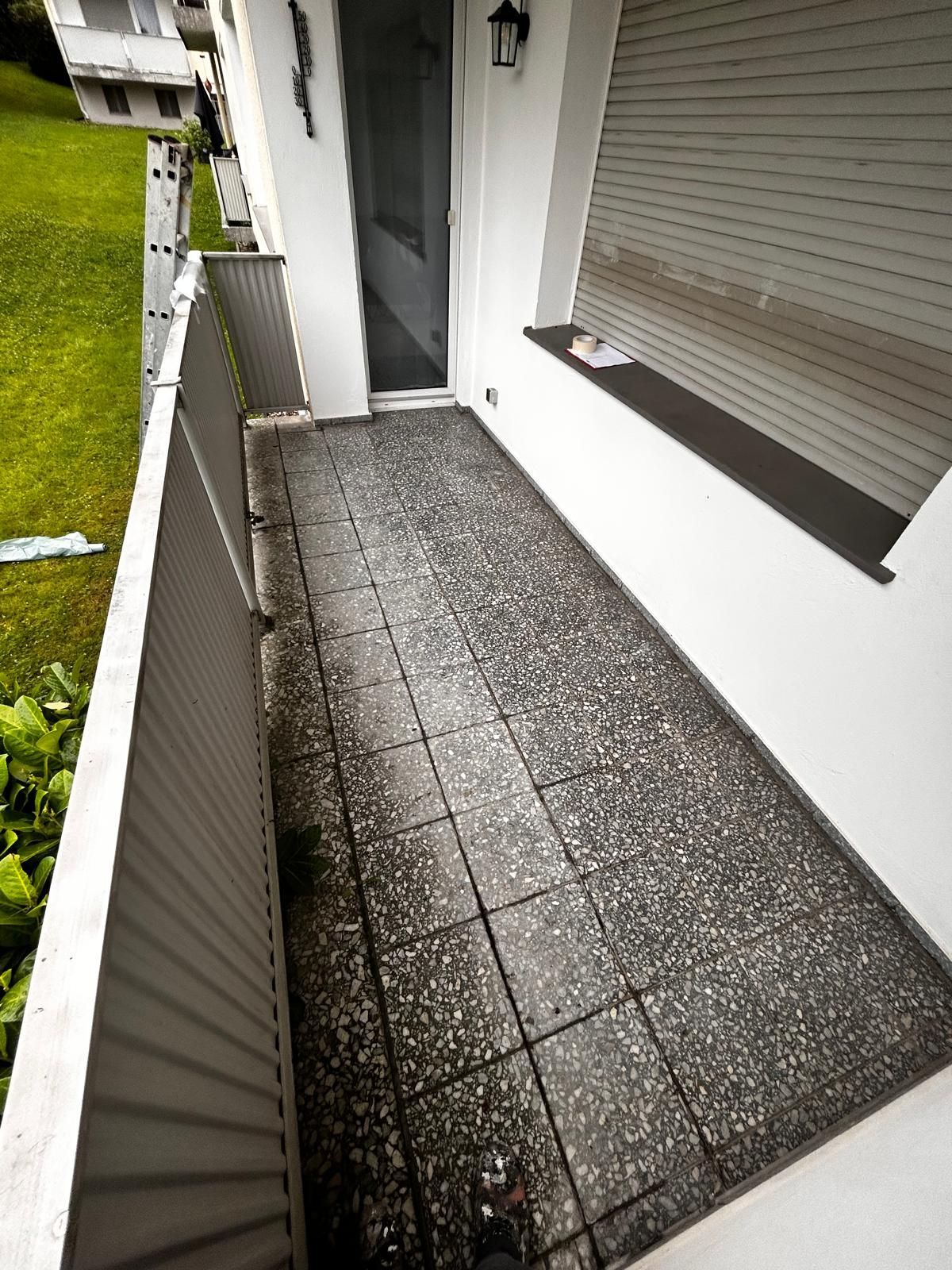 Balkon mit grauem Fliesenboden, weißen Wänden und Metallgeländer. Tür zur Wohnung offen, mit Blick auf grünes Gras.