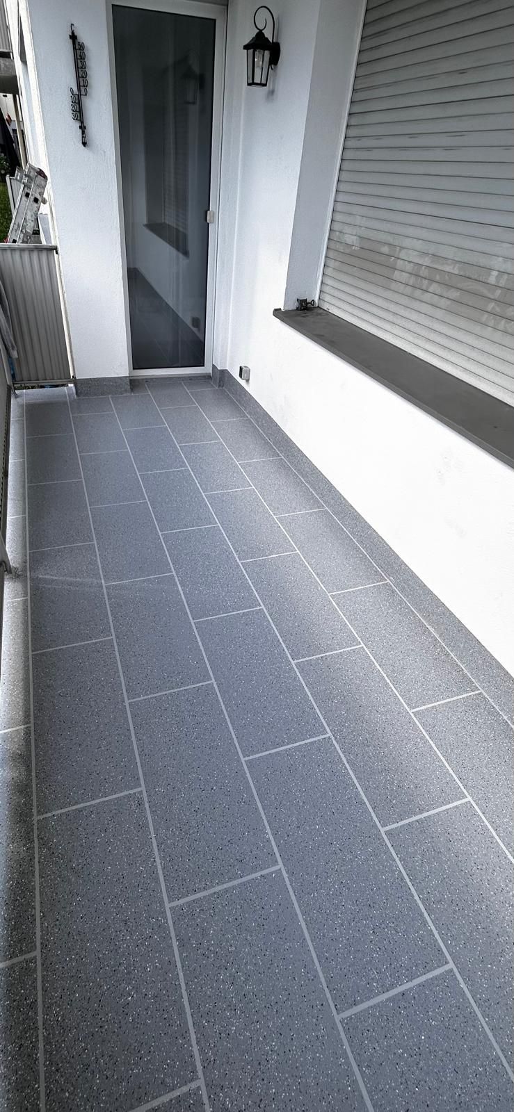 Eine schmale Veranda mit grauem Fliesenboden, weißen Wänden und einer dunkelgrauen Tür.