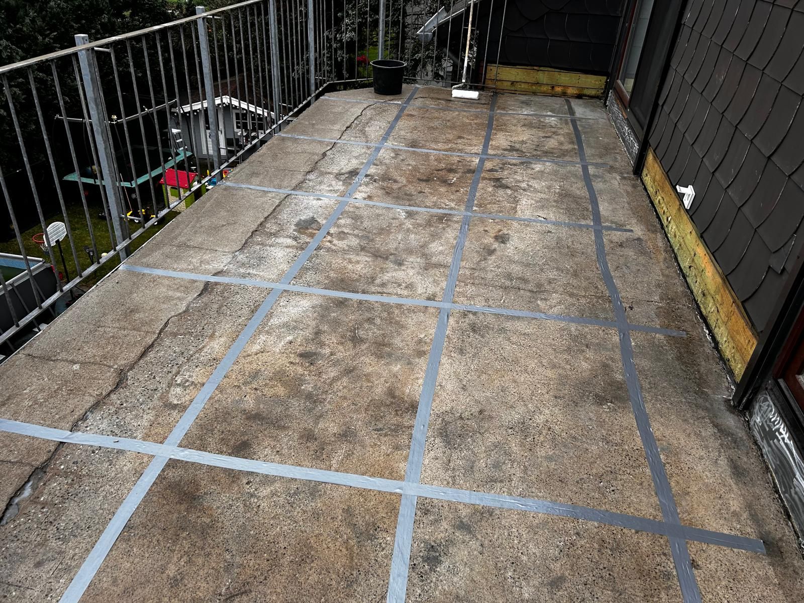 Betonbalkon mit grauem Bandgitter, einem schwarzen Eimer und einer Rolle. Geländer links.