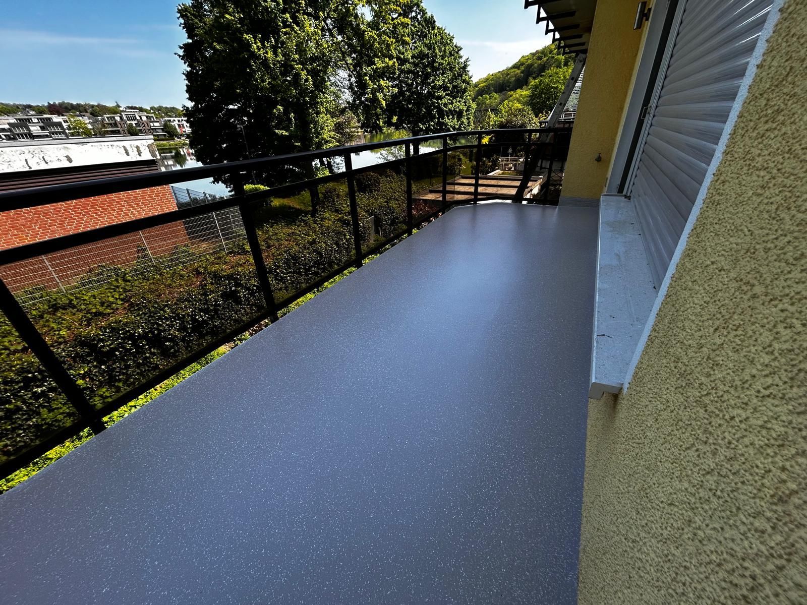 Balkon mit grau gesprenkeltem Boden, schwarzem Geländer und Blick auf Bäume und Gebäude.