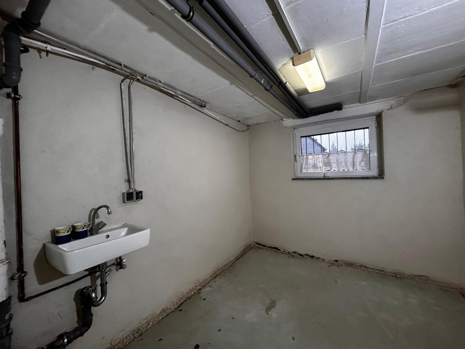 Kellerraum mit Waschbecken, Fenster, Rohren und Leuchtstoffröhre. Betonboden und weiße Wände.
