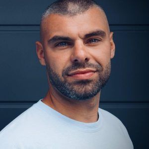 Ein Mann mit rasiertem Kopf, grauem Bart und blauen Augen, der ein weißes Hemd trägt, blickt den Betrachter direkt an.