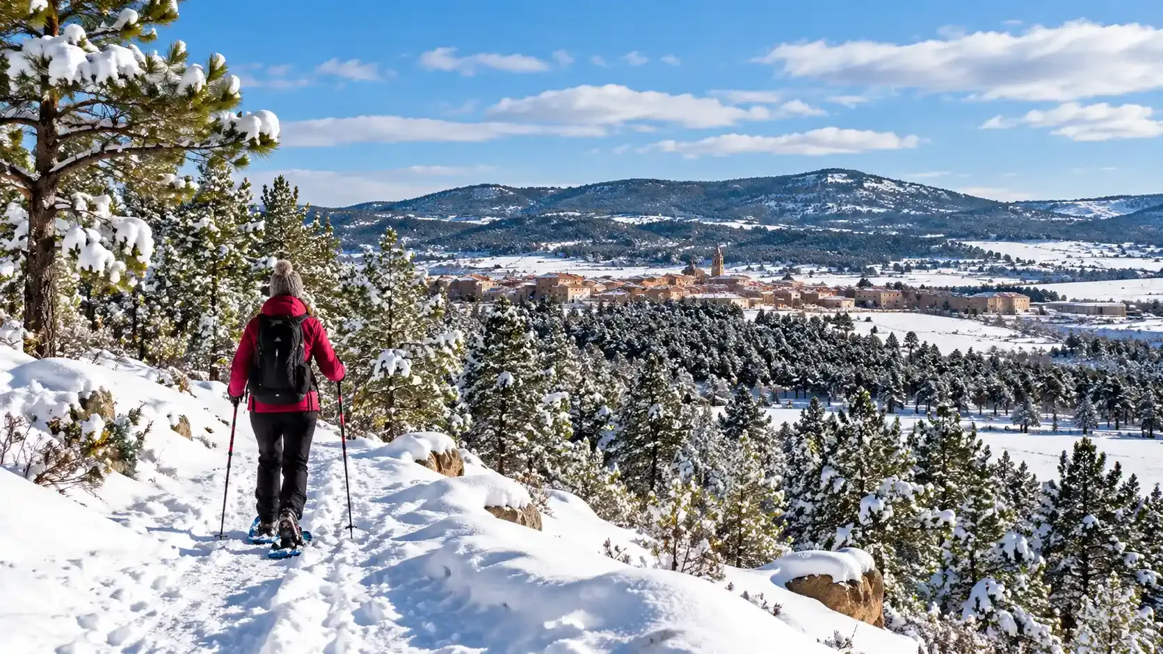 Senderista con una chaqueta roja recorriendo un sendero nevado con vistas a un valle cubierto de pinos y montañas lejanas