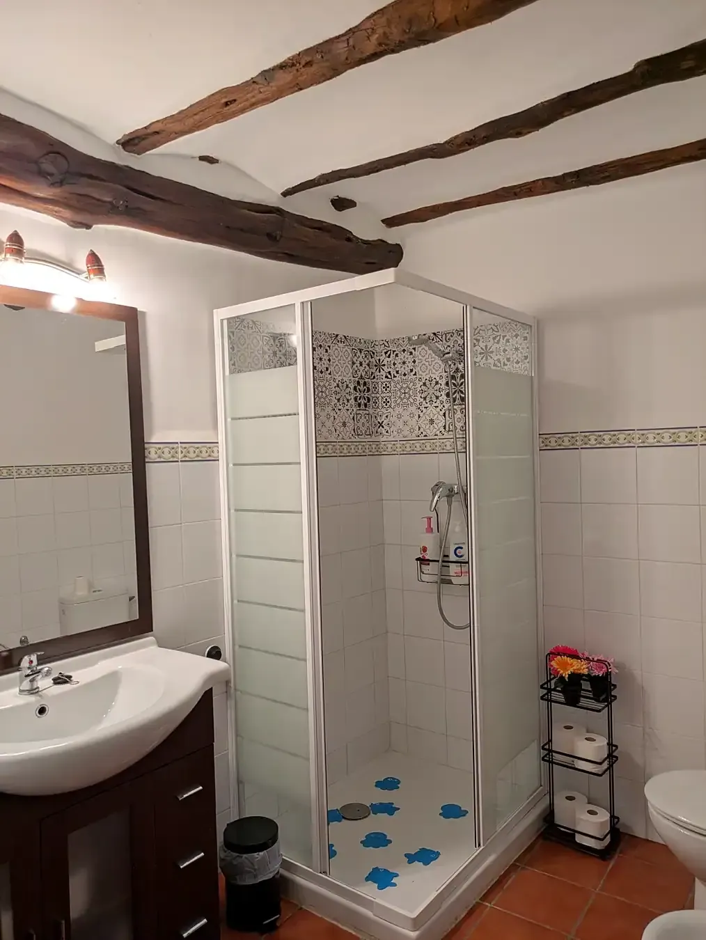 Baño con ducha alicatada en blanco, lavabo, espejo y vigas de madera vistas en el techo.