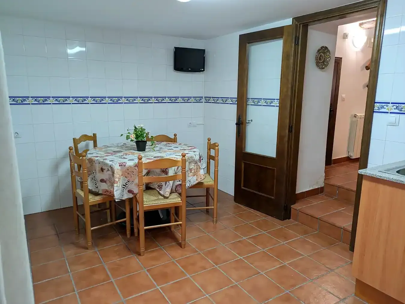 Cocina pequeña con una mesa de comedor de madera, sillas, suelo de baldosas y una puerta abierta que da a un pasillo.