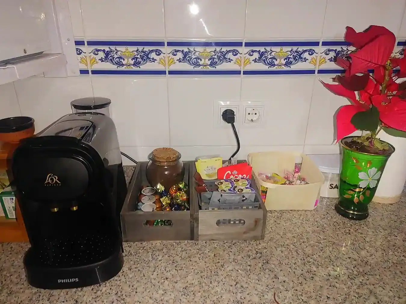 Encimera de cocina con una cafetera negra, recipientes pequeños, un cuenco y una flor de Pascua roja en un jarrón verde.