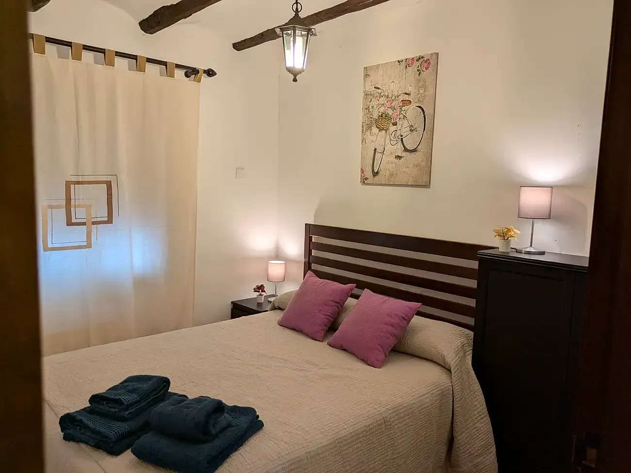 Dormitorio acogedor con colcha beige, almohadas moradas, lámparas de mesita de noche, cortinas y cuadros en la pared