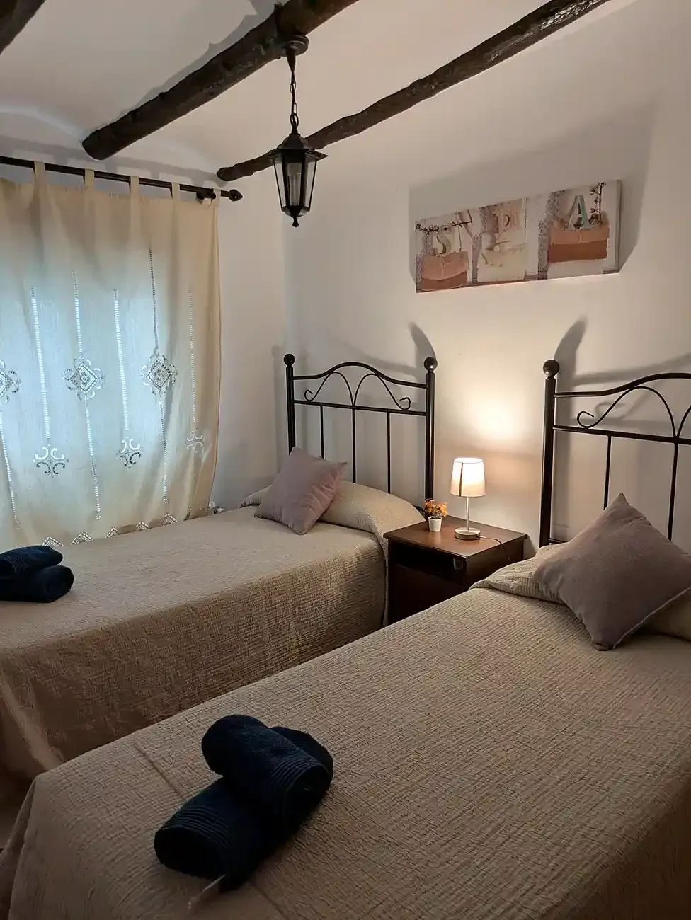 Acogedora habitación doble con camas de hierro forjado, ropa de cama beige, lámpara de mesilla y cortinas de gasa