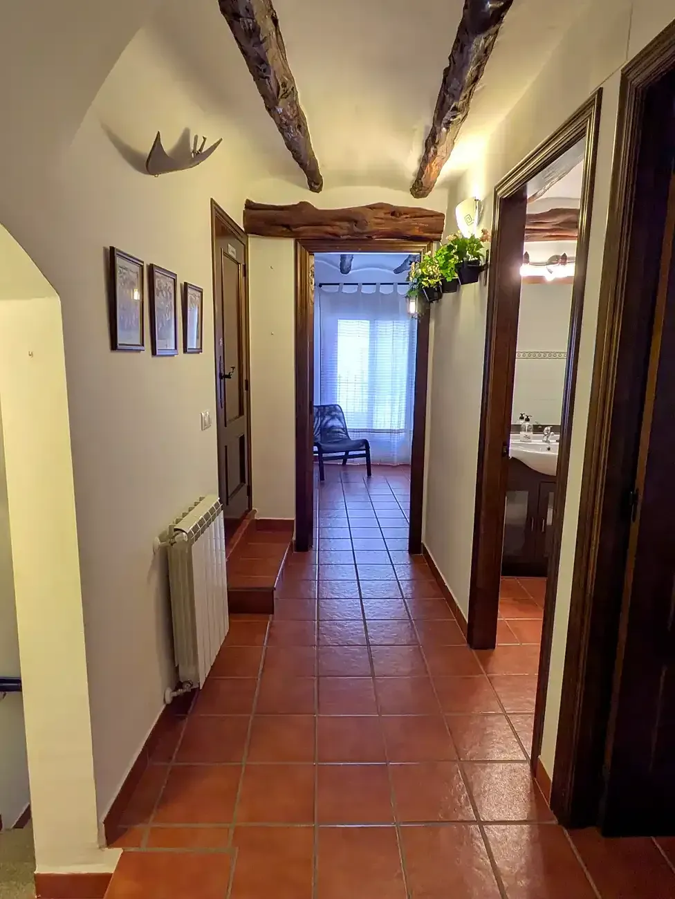 Pasillo estrecho con suelo de baldosas de terracota, vigas de madera a la vista y una puerta luminosa al fondo.
