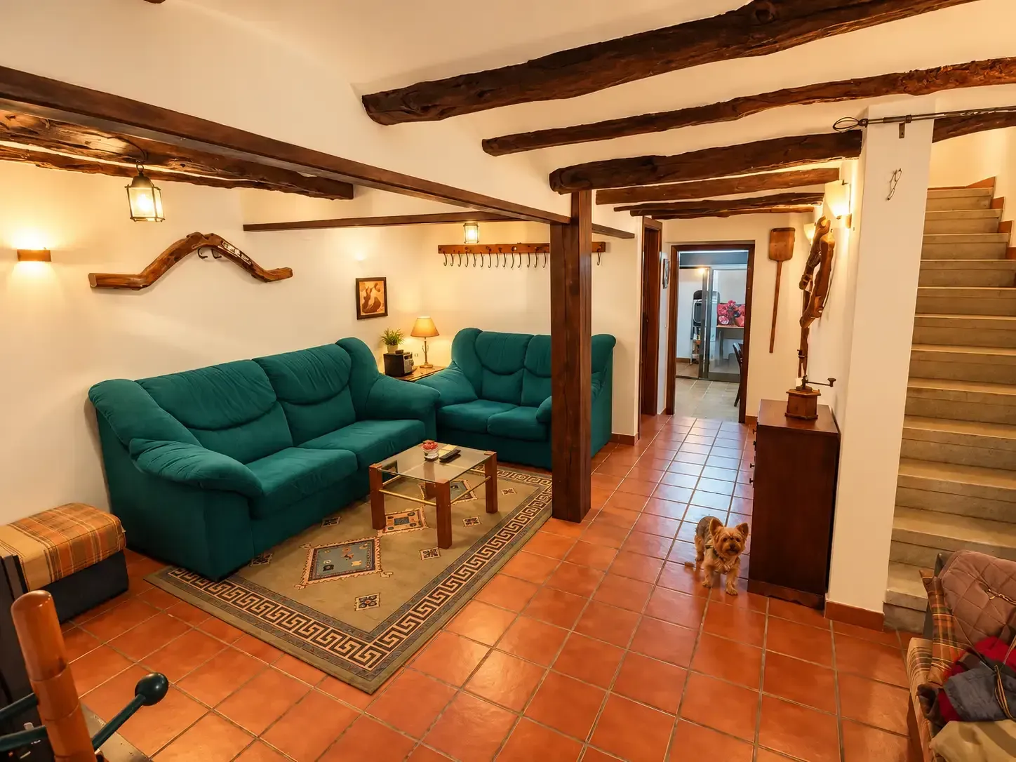 Acogedora sala de estar con sofá verde azulado, suelo de baldosas de terracota, vigas vistas y escaleras a la derecha.