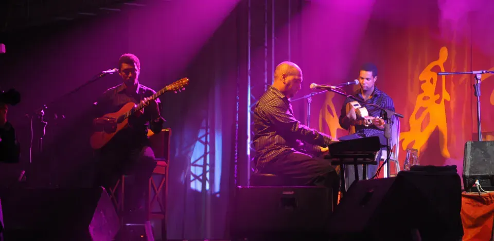 Três homens tocam instrumentos em um palco com luzes roxas atrás deles.
