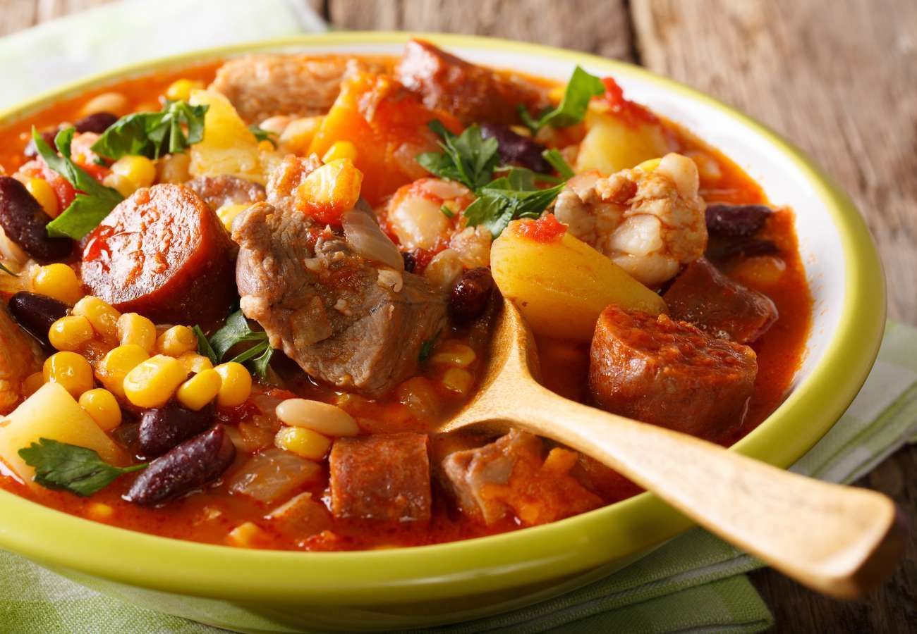 A bowl of soup with meat , beans , corn and sausage with a wooden spoon in it.