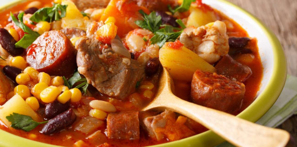 Close-up de um ensopado substancioso em uma tigela verde, com pedaços de carne, linguiça, feijão, milho, batatas e salsa.