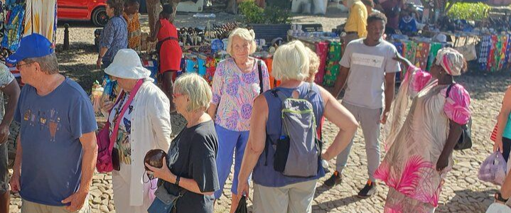 Pessoas caminhando e interagindo em um mercado ao ar livre, com vendedores e tecidos coloridos.