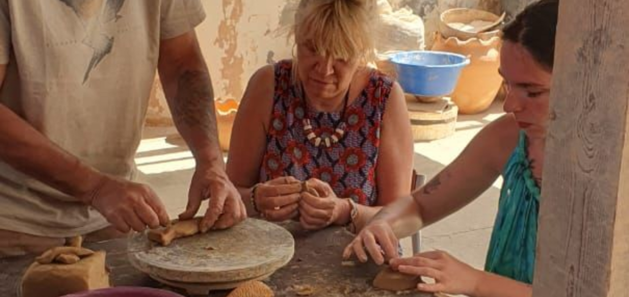 Três pessoas trabalhando com argila em uma mesa, provavelmente cerâmica. Mãos moldando a argila, ambiente externo ensolarado.