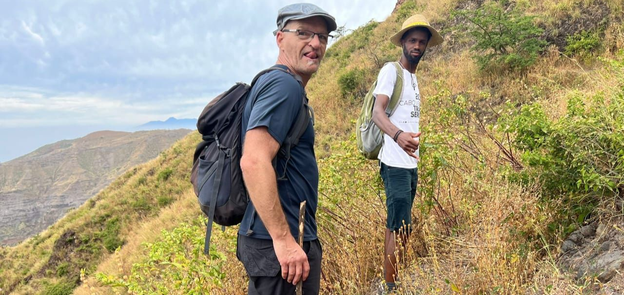 Dois homens caminhando por uma trilha na montanha. Um deles olha para a câmera. Encosta seca e gramada, com céu nublado.
