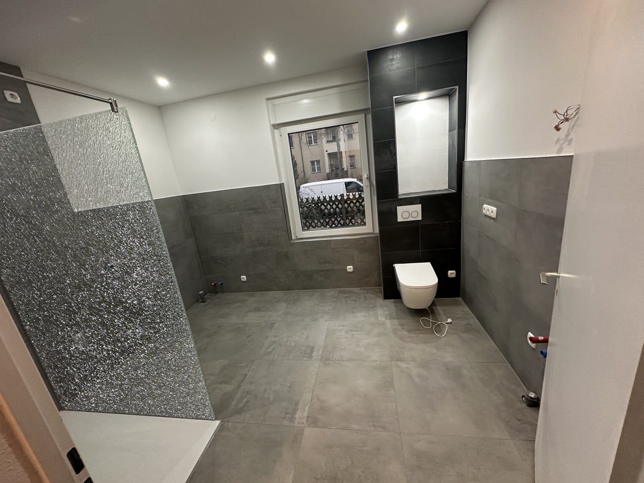 Modernes Badezimmer mit grau gefliesten Böden und Wänden. Dusche, Toilette und Fenster sichtbar.