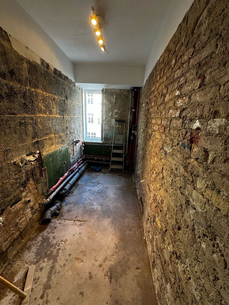 Schmaler Raum mit unverputzten Ziegelwänden, der gerade renoviert wird. Fenster am anderen Ende, Rohre entlang des Bodens.