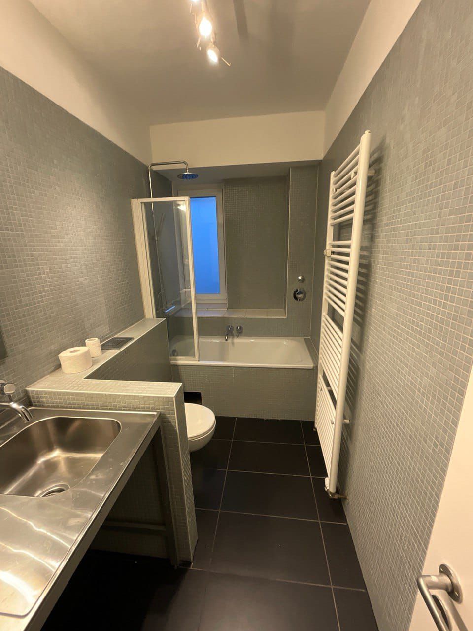 Modernes Badezimmer mit Badewanne, Dusche, Toilette und Waschbecken aus Edelstahl. Wände aus grauen Mosaikfliesen und Boden aus schwarzen Fliesen.