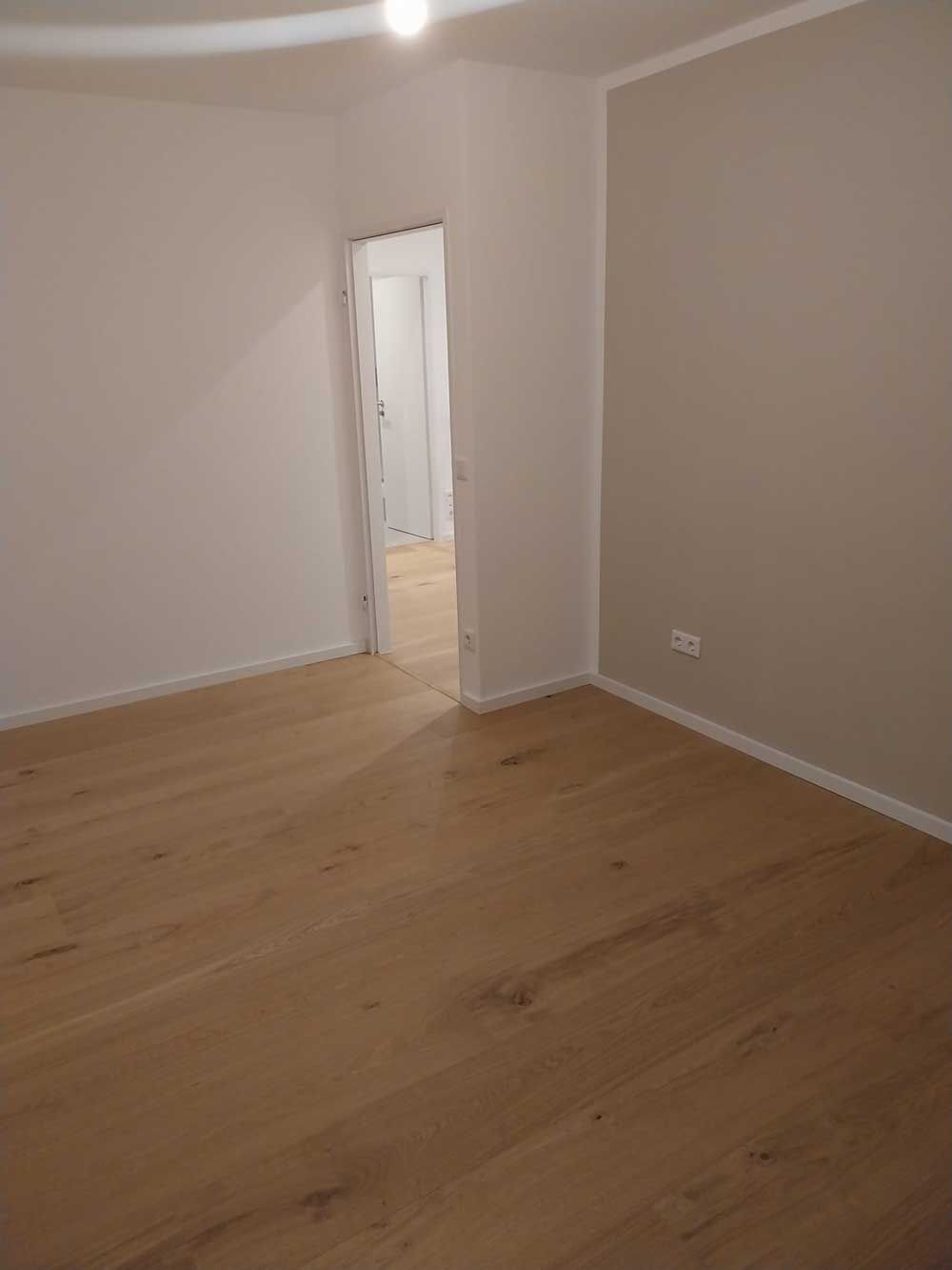 Leerer Raum mit hellem Holzboden, weißen und beigen Wänden und einer offenen Tür.