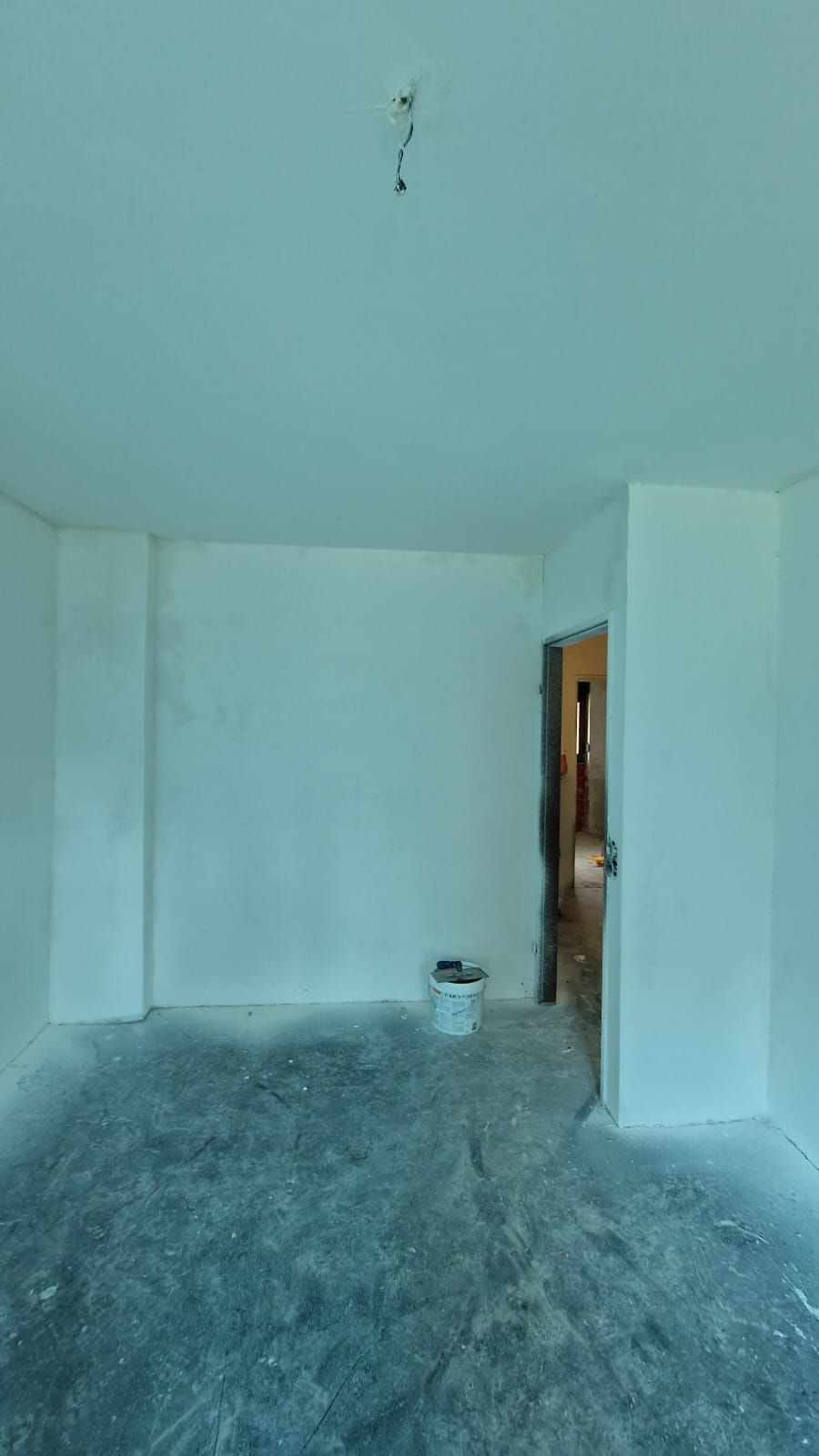 Leerer Raum mit weißen Wänden, unfertigem Betonboden und Türrahmen. Auf dem Boden steht ein Eimer.