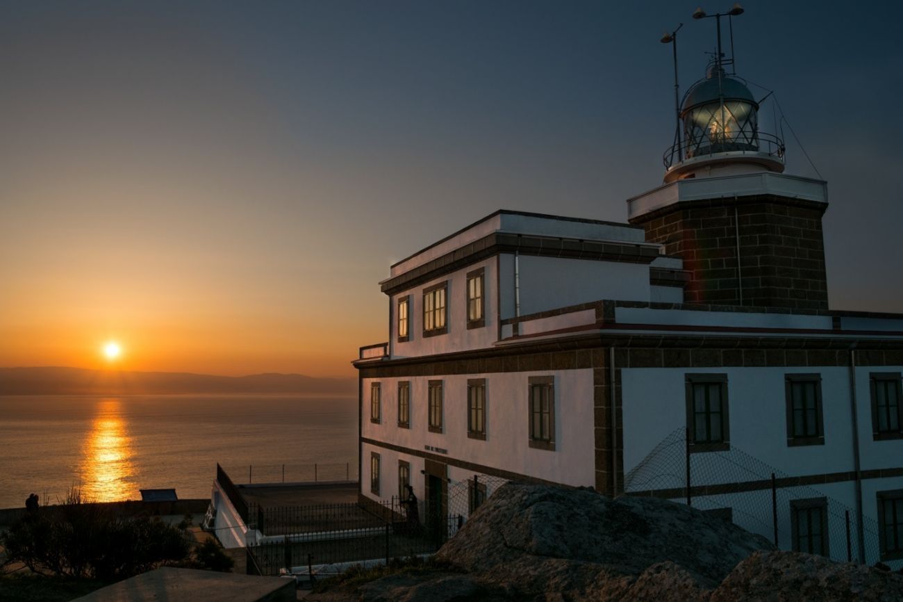 Faro al atardecer, su luz iluminando el agua. Edificio blanco con una torre alta, sol en el horizonte.