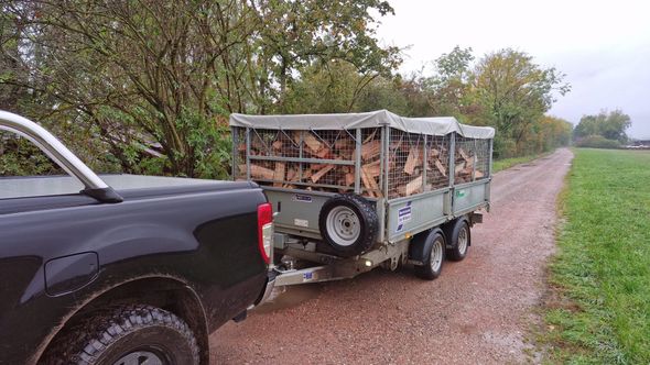 Une camionnette tractant une remorque chargée de bois de chauffage circule sur un chemin de gravier bordant un champ herbeux.