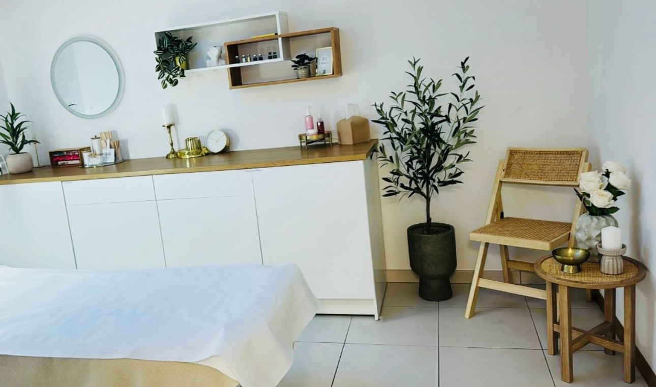 Salle de massage avec armoires blanches, miroir rond, plantes, chaise en bois et table de massage.