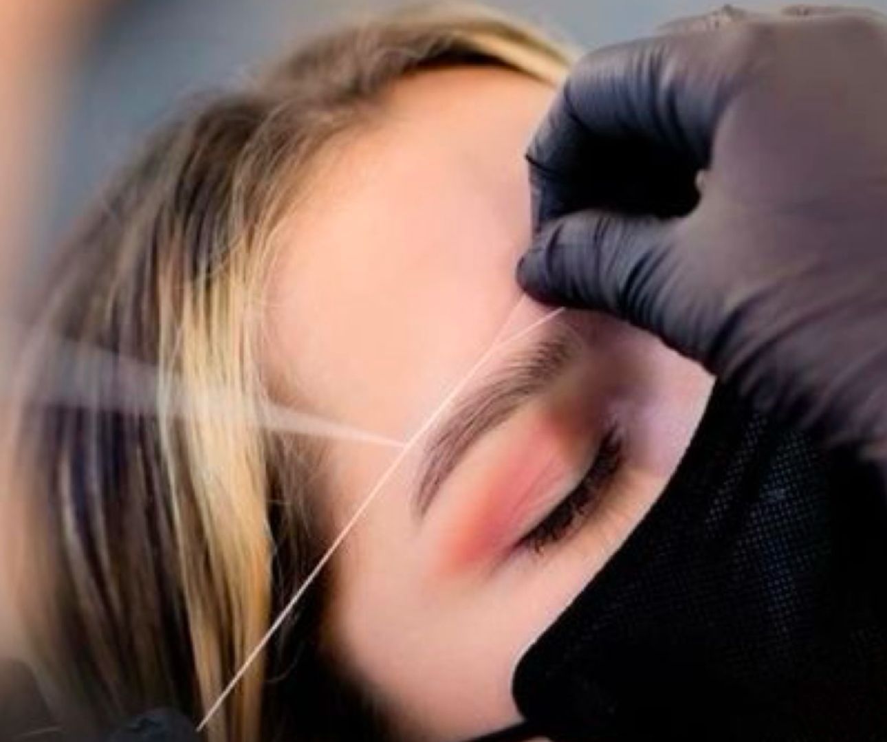 Una mujer se está depilando las cejas con un hilo que lleva guantes negros.