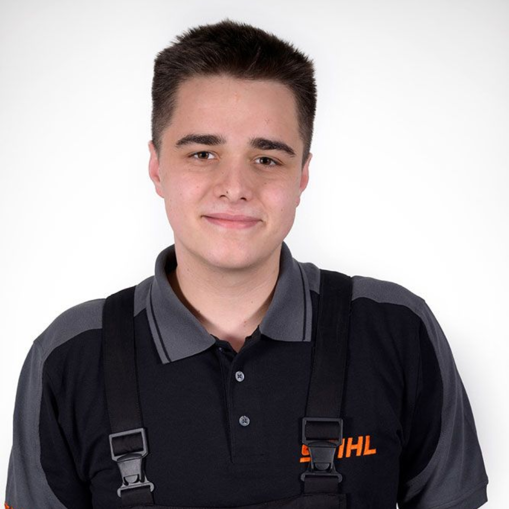 Mann in Arbeitskleidung, lächelnd. Schwarzes Hemd mit grauem Kragen und Latzhose, weißer Hintergrund.