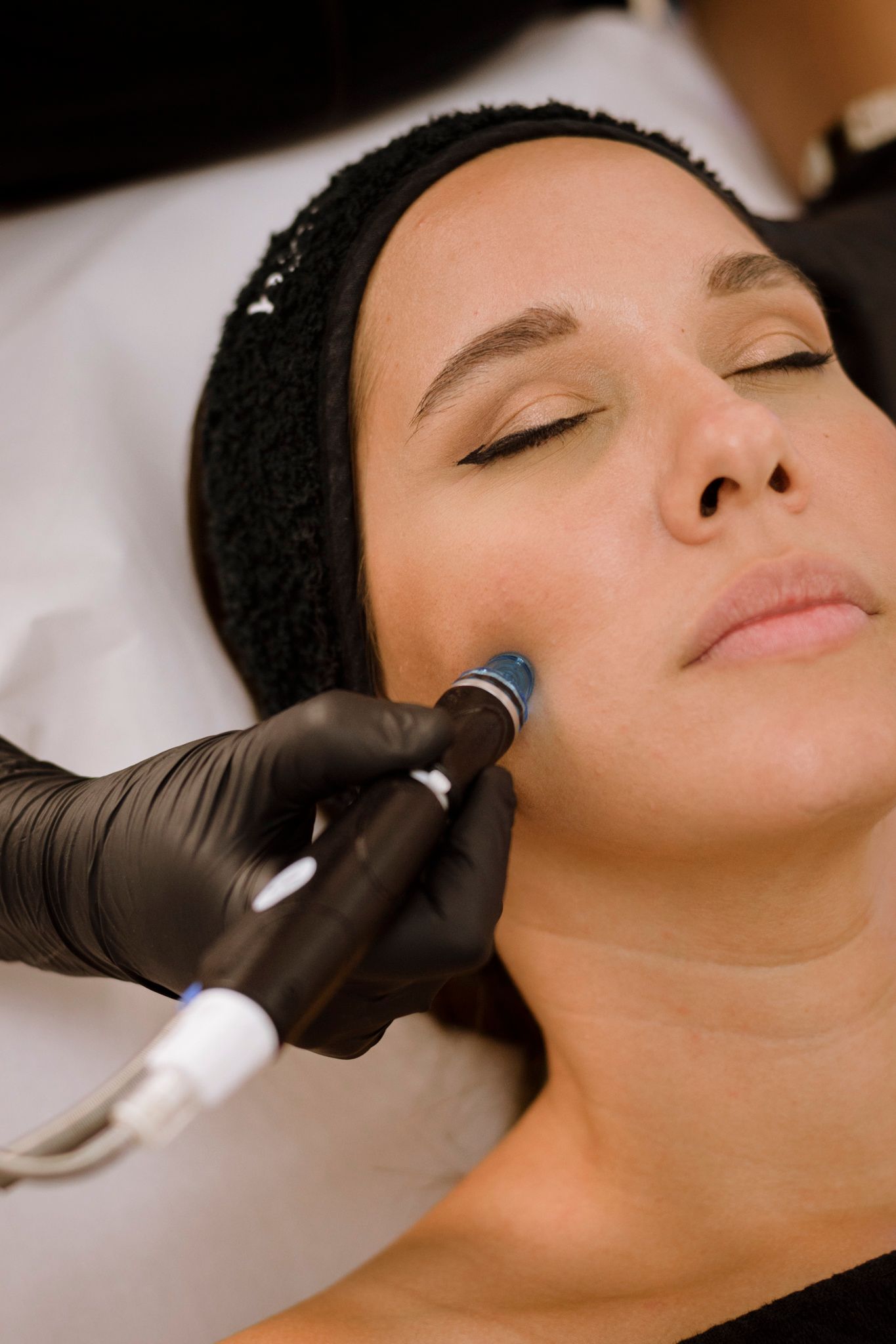 Mujer recibe un tratamiento facial con una mano que lleva un guante negro, en un entorno de spa.