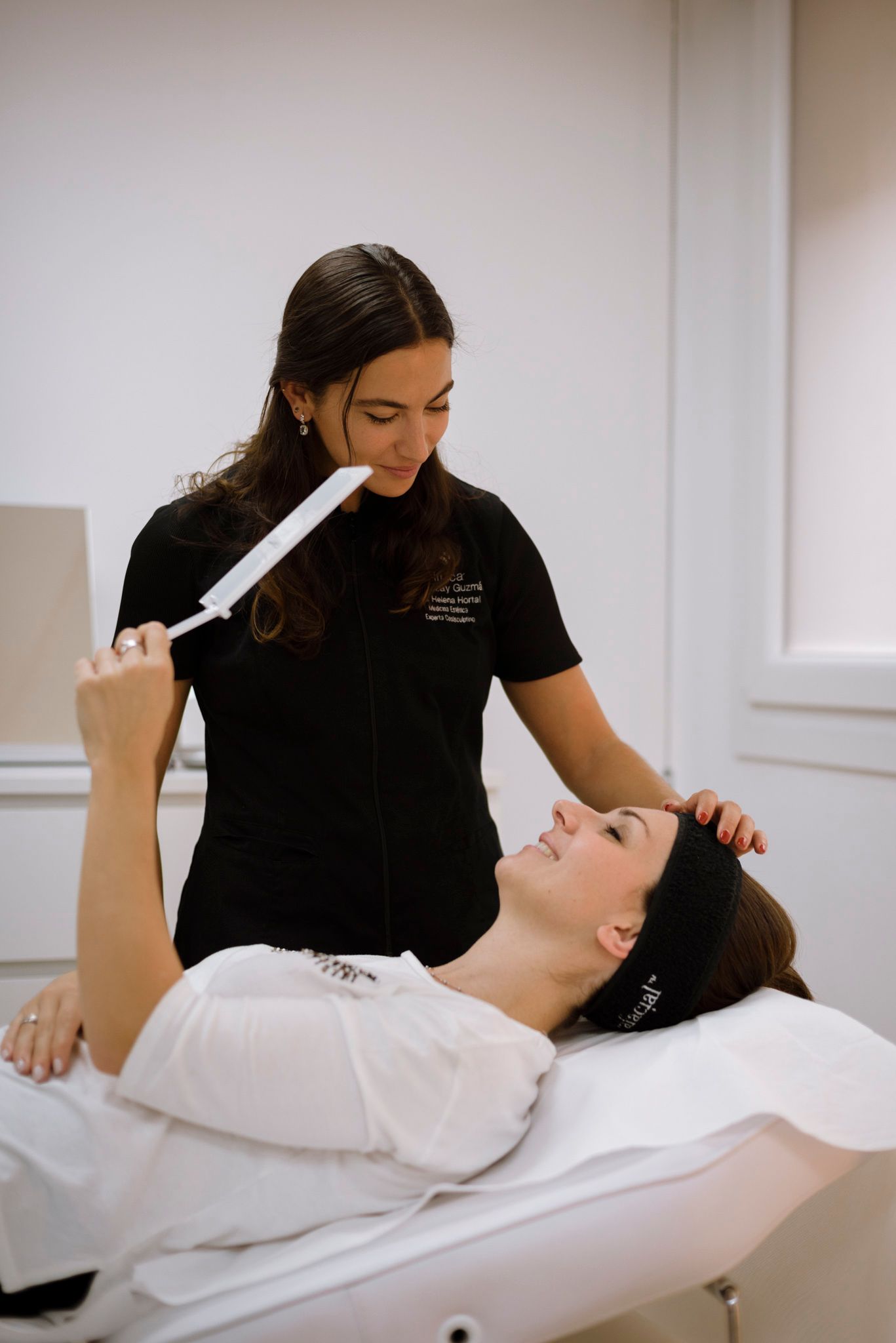 Mujer recibe tratamiento, acostada en una mesa, mientras otra mujer sostiene un dispositivo sobre su cara.