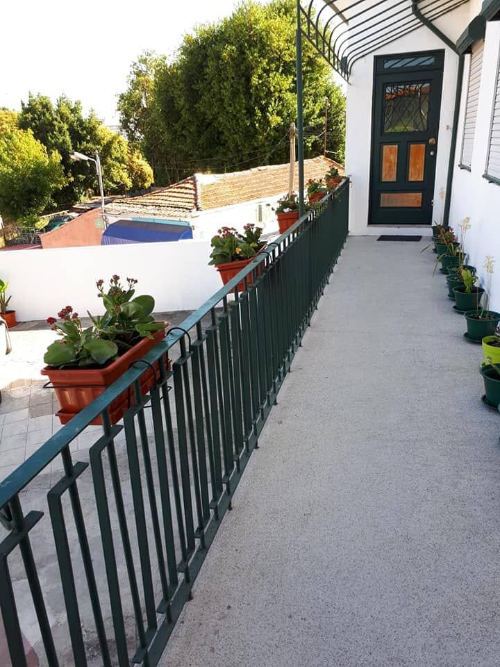 Corrimão verde ao longo de uma passarela de concreto com vasos de flores, que leva a uma porta escura.