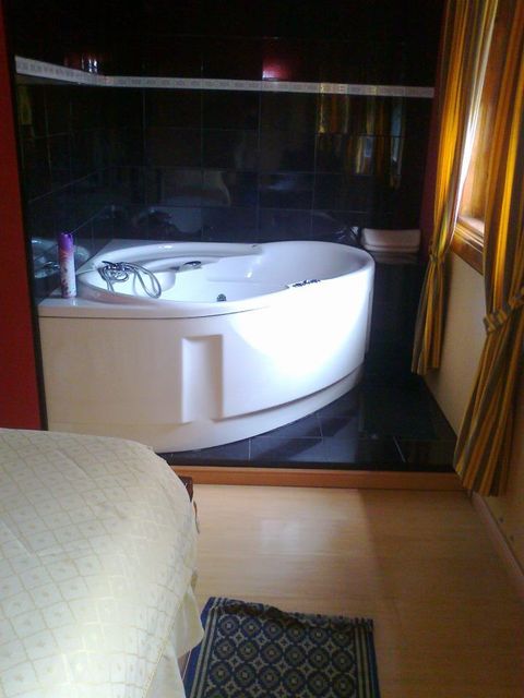 Uma banheira de hidromassagem branca em um banheiro com azulejos pretos, ao lado de uma cama .
