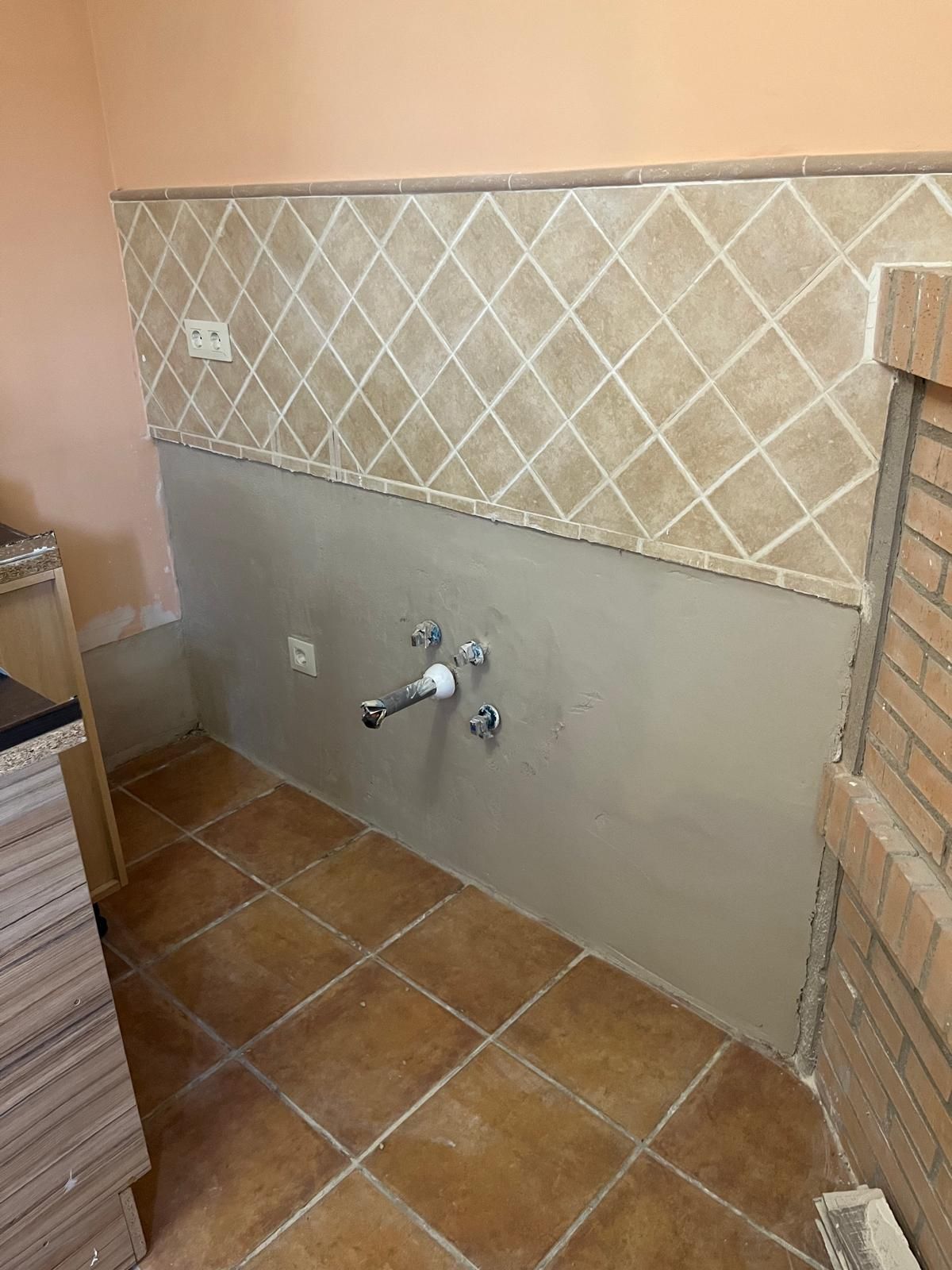 Una pared de cocina con azulejos de color beige con diseño de rombos sobre una sección lisa y sin acabado de color gris