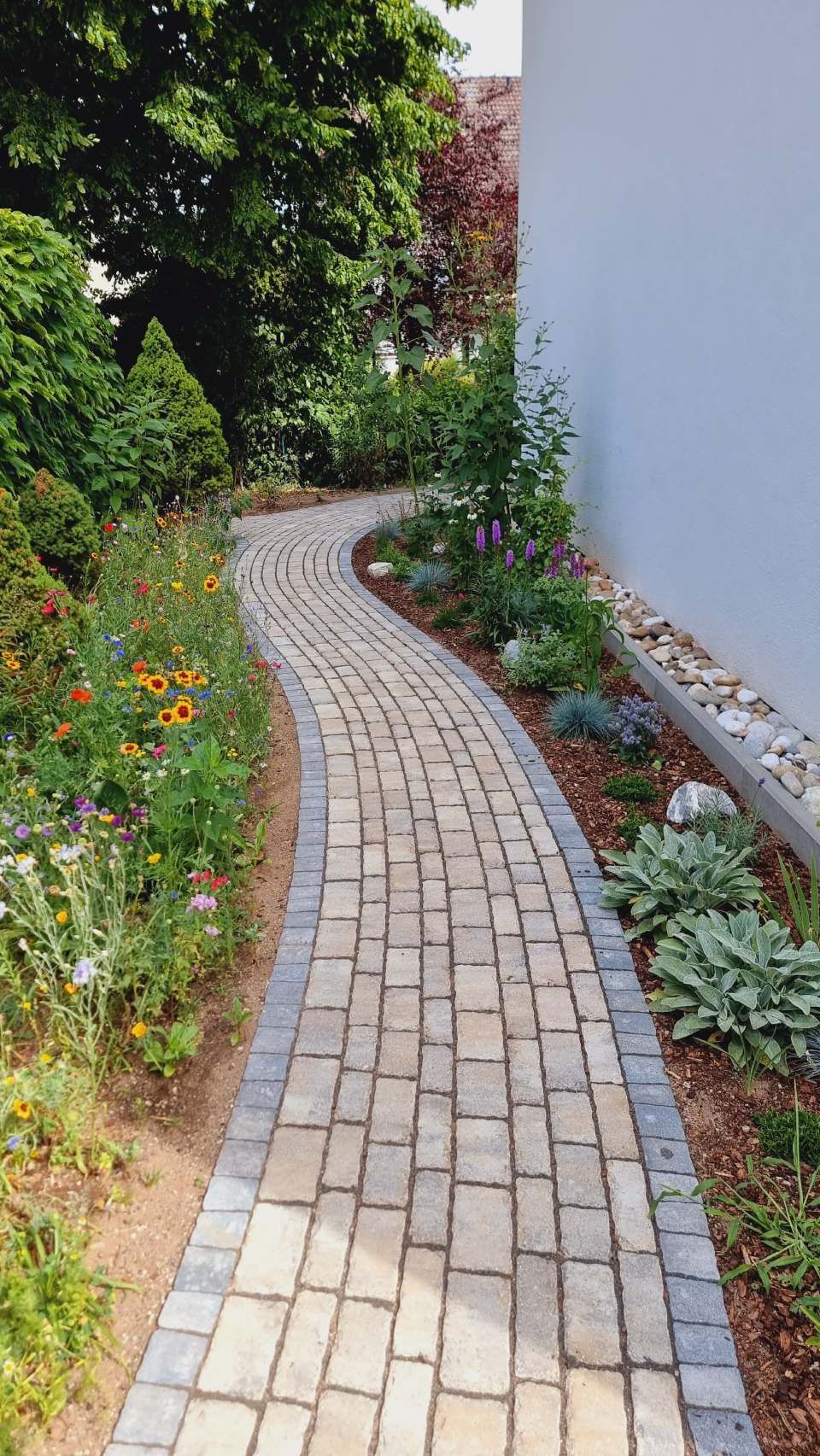 Geschwungener Backsteingartenweg neben einer weißen Mauer, gesäumt von üppigem Grün und farbenfrohen Blumen.