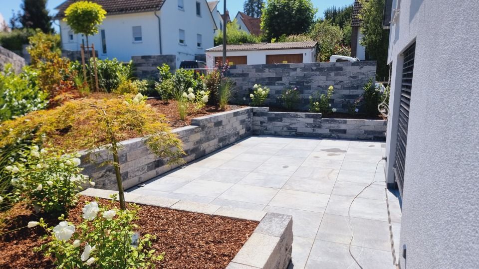 Hinterhofterrasse mit hellen Steinplatten, Hochbeeten und einem kleinen Baum im hellen Sonnenlicht